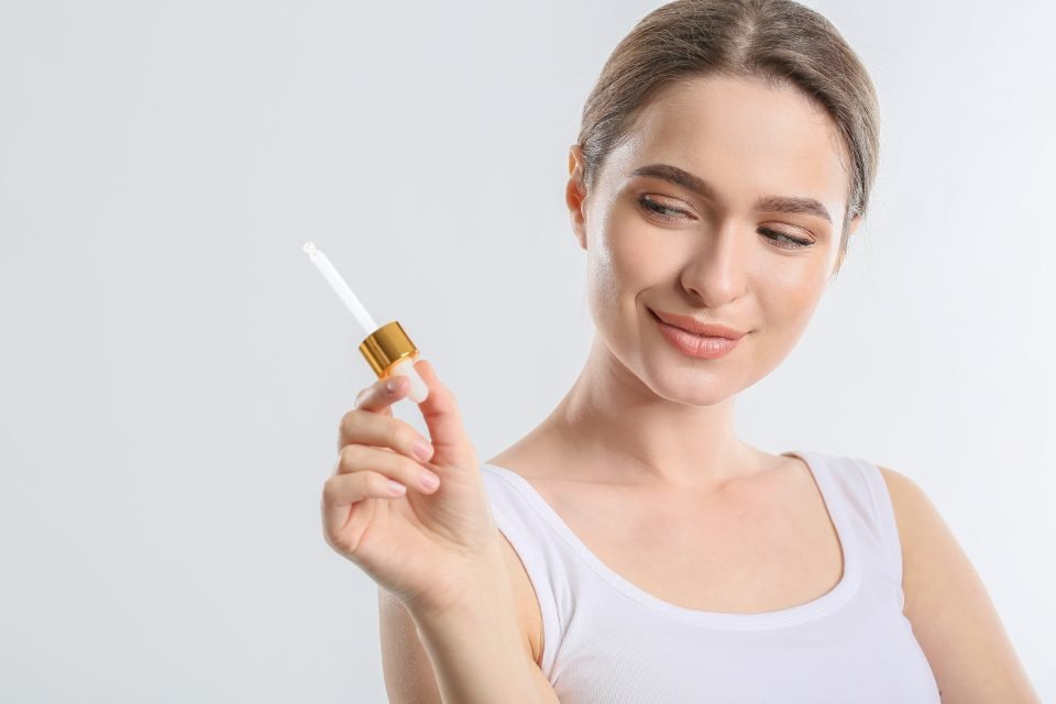 Frau im weißen Top hält lächelnd eine Pipette mit Goldkappe vor hellem Hintergrund