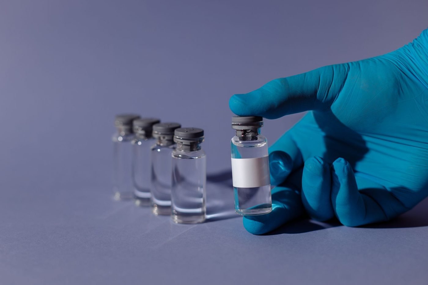 Hand im blauen Einmalhandschuh hält eine unbeschriftete Glasampulle mit klarer Flüssigkeit, dahinter vier weitere