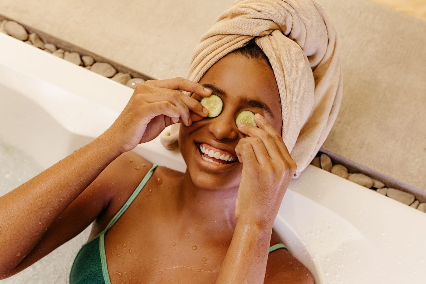 Lachende Frau mit Handtuch-Turban hält sich in der Badewanne zwei Gurkenscheiben vor die Augen